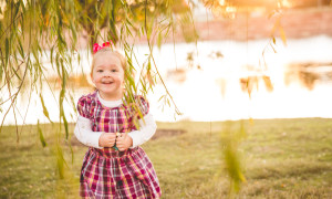 little girl under willow tree in front of sunset lake